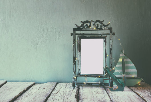 low key image of old victorian steel blue blank frame with fairy garland lights and wooden sailing boat on wooden table. retro filtered image