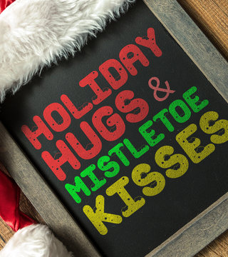 Holiday Hugs & Mistletoe Kisses written on blackboard with santa hat