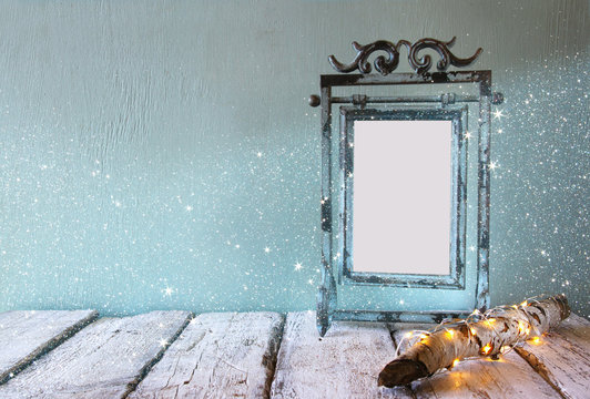 low key image of old victorian steel blue blank frame next to log tree with fairy christmas lights on wooden table

