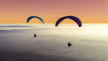Twee paragliders boven een zee van wolken met een pastelkleurige lucht