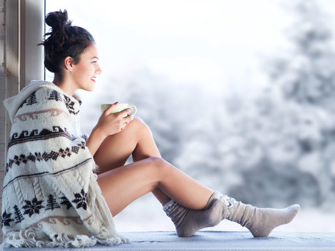 Young beautiful brunette woman drinking cup of coffee wearing knitted nordic print poncho sitting home by the window. Blurred winter snow tree background