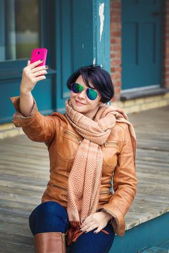 Portrait of middle aged caucasian brunette woman with short bob hair  in sunglasses wearing light brown leather jacket and scarf outside making selfie photo with her phone