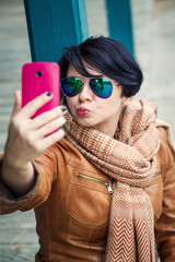 Portrait of middle aged caucasian brunette woman with short bob hair  in sunglasses wearing light brown leather jacket and scarf outside making selfie photo with her phone