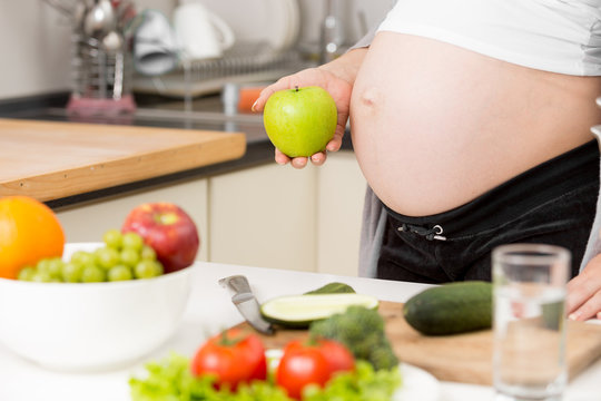 Closeup of pregnant woman posing with green apple on kitchen