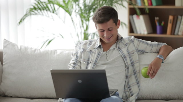 Attractive guy sitting on sofa using laptop eating apple and smiling at camera