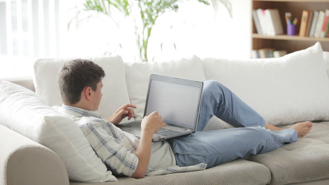 Young man lying on sofa working on laptop looking at camera and smiling