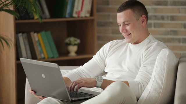 Handsome guy sitting on sofa using laptop and smiling at camera