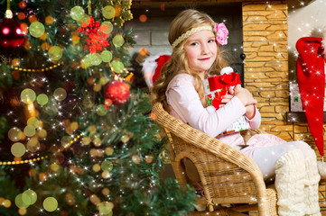 Happy cute toddler girl with beautiful flower in hair sitting under xmas tree with box of gift presents ready to make her wish and celebrate Christmas and New Year