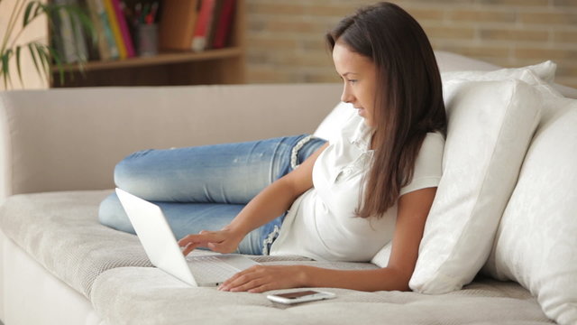 Cheerful girl relaxing on sofa using laptop looking at camera and smiling