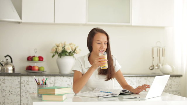 Cheerful student girl sitting at table using laptop drinking juice and smiling