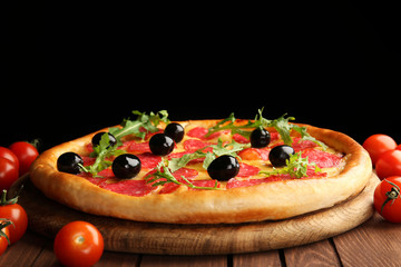 Tasty pizza with salami and olives decorated with tomatoes on wooden table against black background