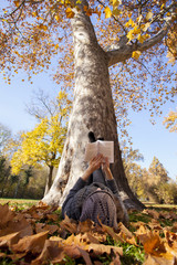 Young girl reading a book lying in the grass on the leaves in the autumn park
