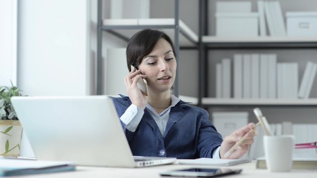 Happy businesswoman sitting at office desk, chatting on the phone and laughing