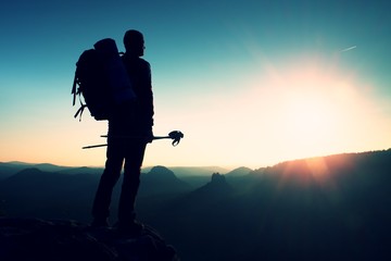 Tall backpacker with poles in hand. Sunny hike day in rocky mountains. Hiker with big backpack stand on rocky view point above misty valley.