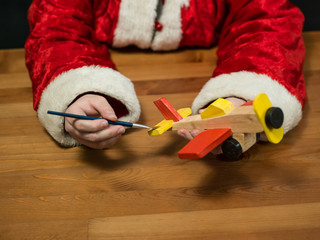 Santa Claus sitting in his workshop painting a toy airplane. Hor