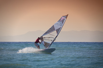 Windsurfen in Rhodos Griechenland. Mann surft im blauen Meer vor rötlichem Himmel, Insel im Hintergrund.
