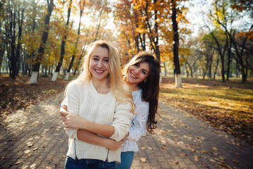 Fototapeta premium Two happy and cheerful young girl students, blond and brunette in coat and jeans laughing in sunny autumn park full of fallen leaves, horizontal pictures