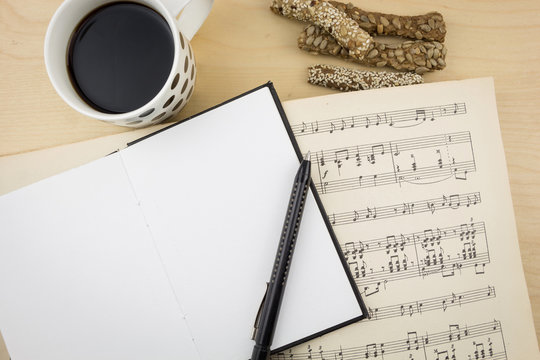 Opened blank notebook with pen, cup of coffee and music notation book, on wooden desktop.
