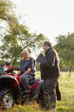 Two men, a farmer and a man with a clipboard, by a quadbike in an orchard. 