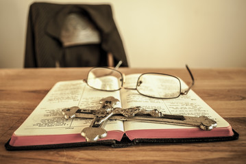 Prayer book with crucifix and glasses. In the bacground an empty chair with man's coat. 