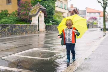 Outdoor portrait of adorable little blond boy with umbrella under the rain, playing in the puddle