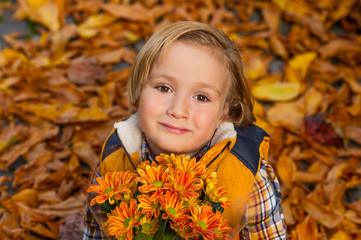 Autumn portrait of adorable little blond boy of 4 years old, wearing warm yellow vest coat, holding small bouquet of orange chrysanthemum flowers