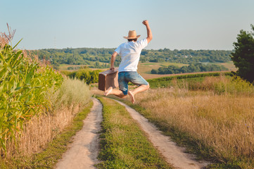 Backview of excited man with suitcase jumping on country road - obrazy, fototapety, plakaty