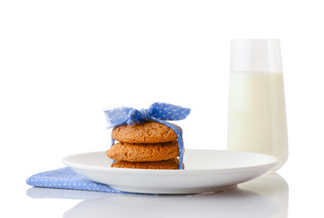 Stack of three homemade oatmeal cookies tied with blue ribbon in small white polka dots on white ceramic plate on matching blue napkin and glass of milk, isolated on white background