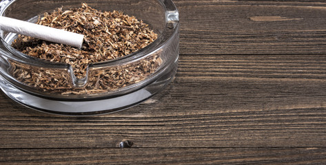 The whole cigarette and a glass ashtray stand on an old wooden table.Crop