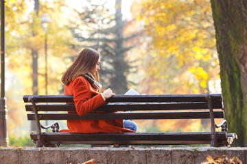 Young girl sitting on a park bench and reading a book, on a beautiful autumn day