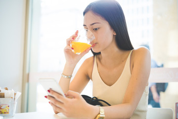 Young handsome asiatic long brown straight hair woman sitting  in a bar, drinking a juice and using a smartphone, looking downward the screen - technology, social network, relaxing concept