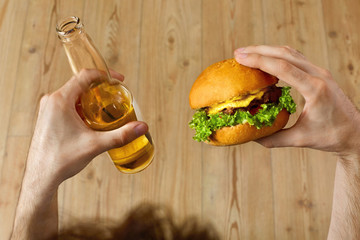 Eating Fast Food. Closeup Of Man's Hands Holding Classic American Hamburger And Bottle Of Beer. Dinner At Restaurant. Point Of View. Nutrition