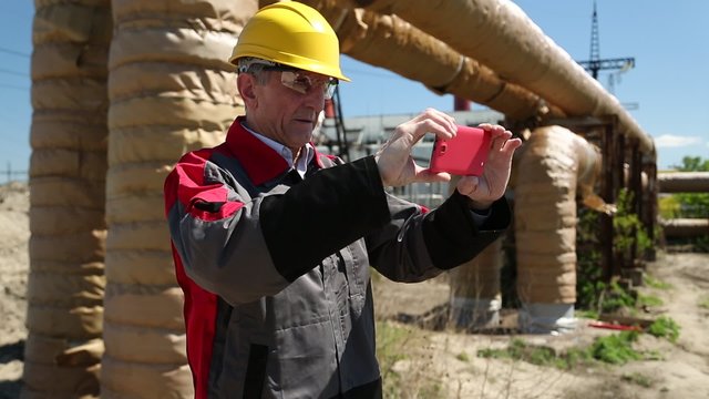 Workman in yellow hard hat at power plant records videos on his smartphone. Supervisory personnel in yellow hard hat with red smartphone near heating main pipeline at heat electric power station
