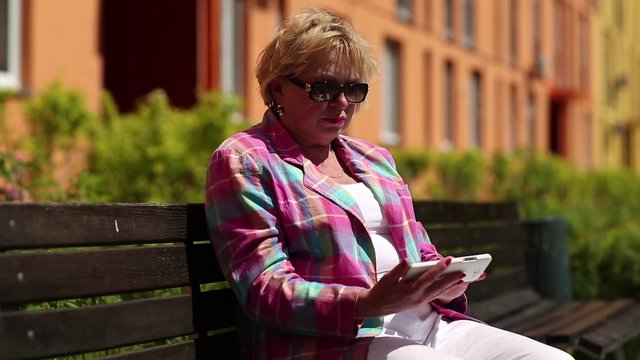 Woman sits on the bench in courtyard and uses white smartphone. Woman looking and flipping through the photos in her cell phone
