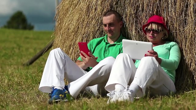 Man with red smartphone and woman with Tablet-PC sits near a haystack. Mother and son with computer and smartphone looks and flips through the photos in their devices
