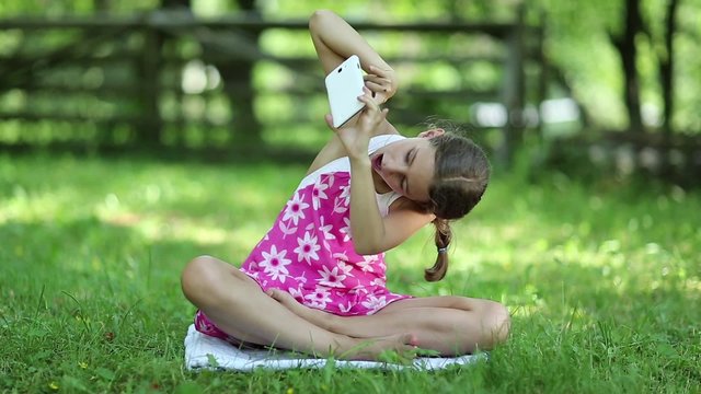 Pretty girl sits on the grass and makes selfie on his smartphone. Beautiful girl in pink dress with mobile phone sits on the grass and makes photos
