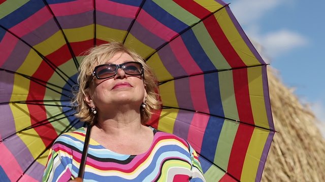 Blonde woman in glasses with brightly coloured umbrella stands near haystack and looks into the distance
