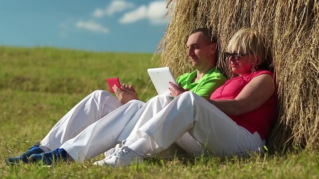 Mother and son with tablet PC and smartphone looks and flips through the photos in their devices. Man with red smartphone and woman with white tablet computer sits near a hayrick

