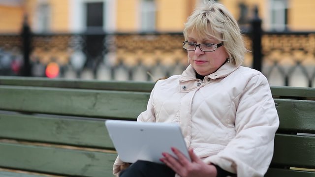 Blonde woman sits on the bench near the road and uses tablet PC. Woman with tablet computer sits on the green bench
