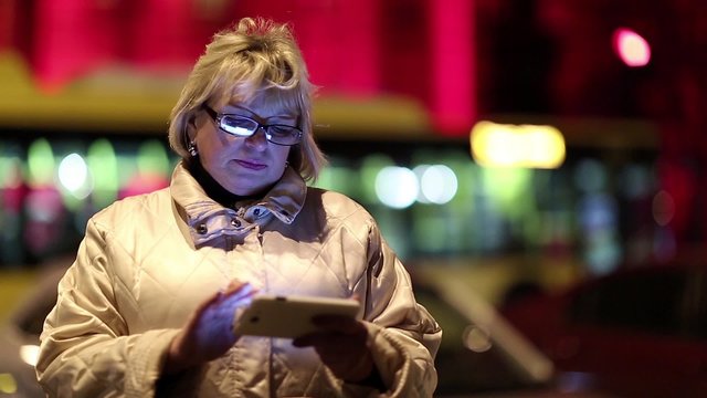Senior woman with cell phone standing near the road in nighttime. Woman with glasses standing near the road and uses smartphone, night city, trolley bus from the background, purple face
