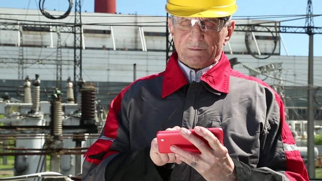 Maintenance worker with red smartphone at power station. Power engineering specialist with red smartphone at heat station. Worker in yellow hard hat at heat station. Factory worker with red smartphone