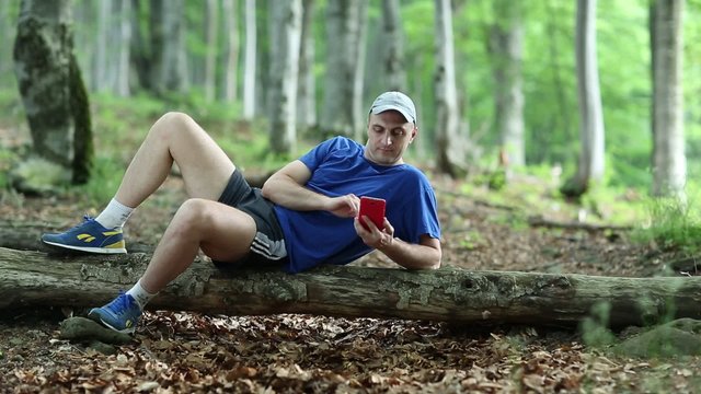 Adult man in dark blue t-shirt with red smartphone lies on a fallen tree in the forest
