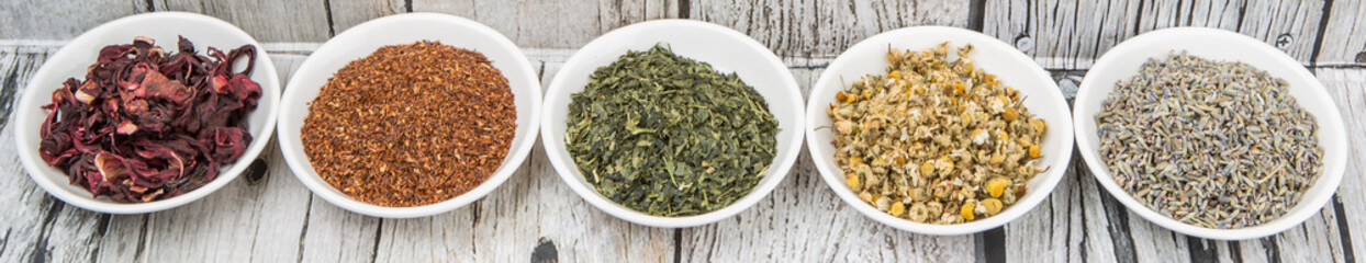 Dried herbal tea leaves, lavender, rooibos, chamomile, linden flower, hibiscus, Japanese green tea in white bowl over wooden background