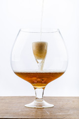 Beer is pouring into a glass which is into another huge glass, creating foam and bubbles on the wooden texture  at the bottom and white background