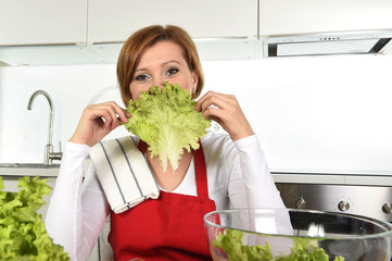 young beautiful woman in red apron at home kitchen preparing vegetable salad bowl playing with with lettuce