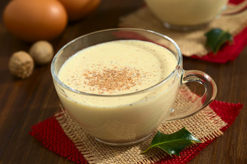 Eggnog drink in glass cup sprinkled with ground nutmeg, photographed on dark wood with natural light (Selective Focus, Focus one third into the eggnog and on the handle of the cup)