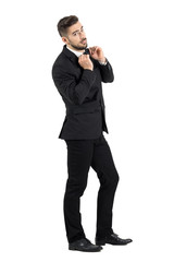 Side view of cool elegant young male model adjusting bow tie looking at camera. Full body length portrait isolated over white studio background.