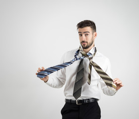 Portrait of a confused young bearded man in white shirt choosing right necktie over gray studio background. 
