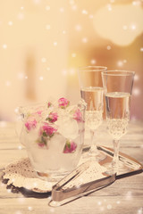 Ice cubes with rose flowers in glass bucket and two glasses with champagne on wooden table, on bright background