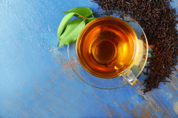 Glass cup of tea with green leaves and scattered tea around on blue wooden background